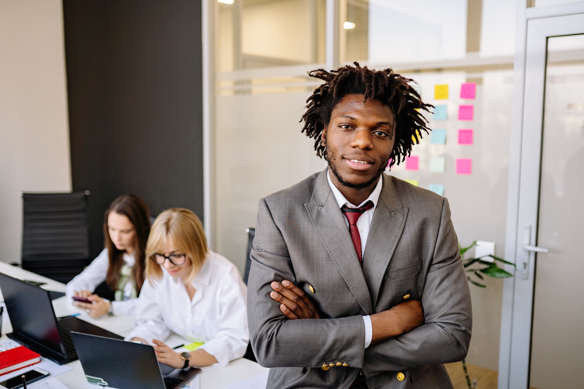 professional-man-suit-arms-crossed Black Hr professional man with arms crossed and female colleagues discussing HR functions behind him